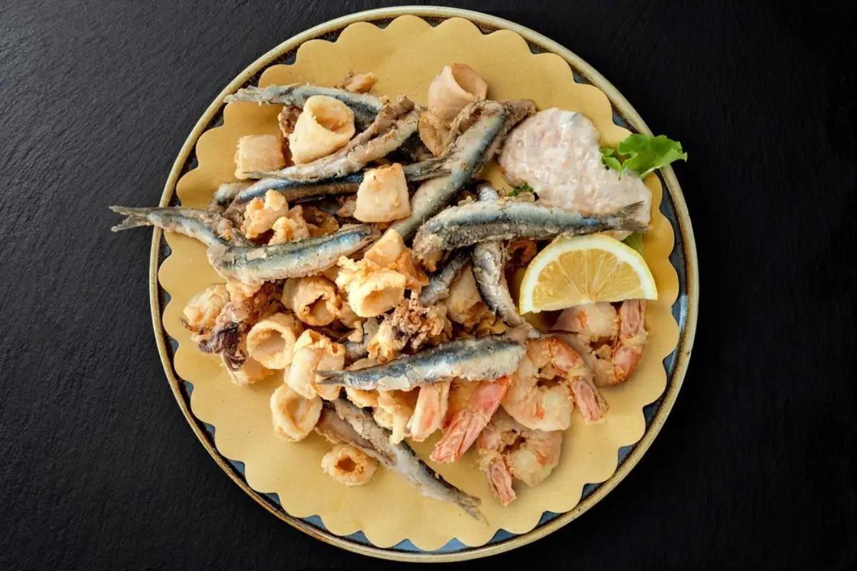 Top view of Round dish with mixed fried fish with sauce and lemon wedge on black stone 
